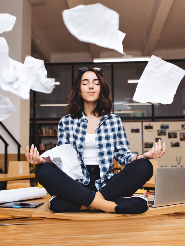 Young pretty joyful brunette woman meditating on table surround work stuff and flying papers. Cheerful mood, taking a break, working, studying, relaxation, true emotions; tips to improve emotional intelligence (EQ) in the workplace