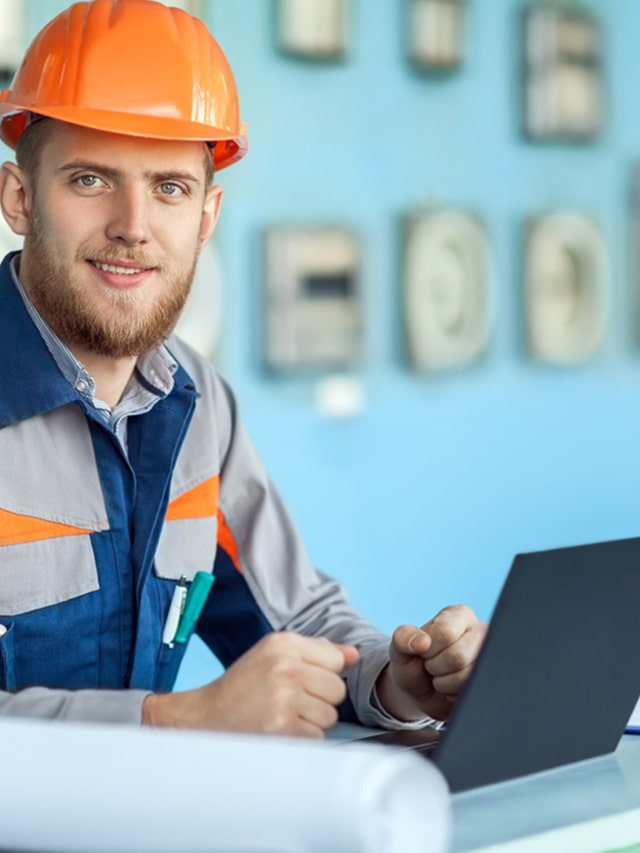 Portrait of young happy engineer working at laptop in control room; concept of Prepare for your electrical engineering job interview with these frequently asked questions.